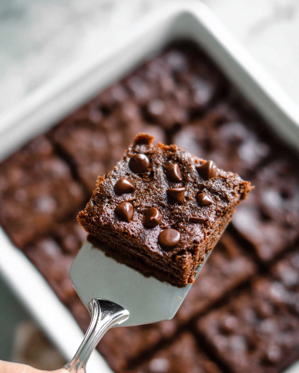 A close-up of a single square piece of chocolate chip brownie being lifted by a woman’s hand holding a silver spatula with a white marbled background below. The brownie piece has one thick, dense dark brown layer with visible moist texture and several shiny, melted chocolate chips on top. Behind it, a white baking pan filled with many more brownies with a missing square piece can be seen slightly out of focus. The whole scene is softly lit. Photo taken with an iphone --ar 4:5 --v 7