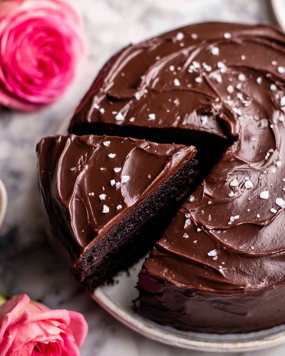A round chocolate cake with a thick, smooth, and glossy dark chocolate frosting covers the top and sides. The top layer of frosting shows swirled patterns with a rich texture. The cake is sliced into three visible pieces, revealing a dark, moist chocolate cake layer underneath. Small flakes of sea salt are sprinkled over the frosting, adding some contrast and texture. The cake sits on a white plate against a white marbled surface, with a bright pink rose placed nearby. Photo taken with an iphone --ar 4:5 --v 7