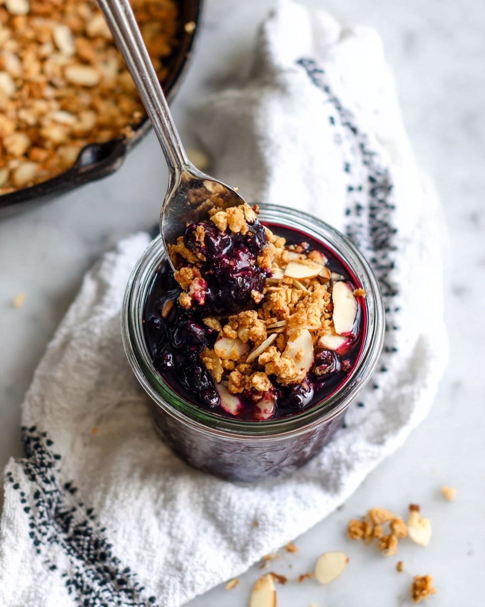The image shows a clear glass jar filled with three visible layers: the bottom and middle layers are dark purple blueberry compote with whole blueberries, rich and shiny, while the top layer is light brown granola with oats and almond slices scattered freely, giving a crunchy texture. A silver spoon is placed in the jar, slightly lifting some granola and compote. The jar sits on a white cloth with black patterns and is on a white marbled surface. Some granola pieces are scattered around the jar. Part of a cast iron pan filled with granola is partially visible in the top left corner. photo taken with an iphone --ar 4:5 --v 7