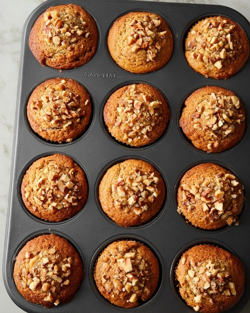 A dark muffin tray holds twelve golden brown muffins, each topped with a generous layer of light brown chopped nuts that add a crunchy texture. The muffins have a slightly domed shape with a soft, moist crumb showing through small cracks on the surface. The tray rests on a white marbled surface, giving a clean and simple background that contrasts with the warm colors of the muffins. photo taken with an iphone --ar 4:5 --v 7