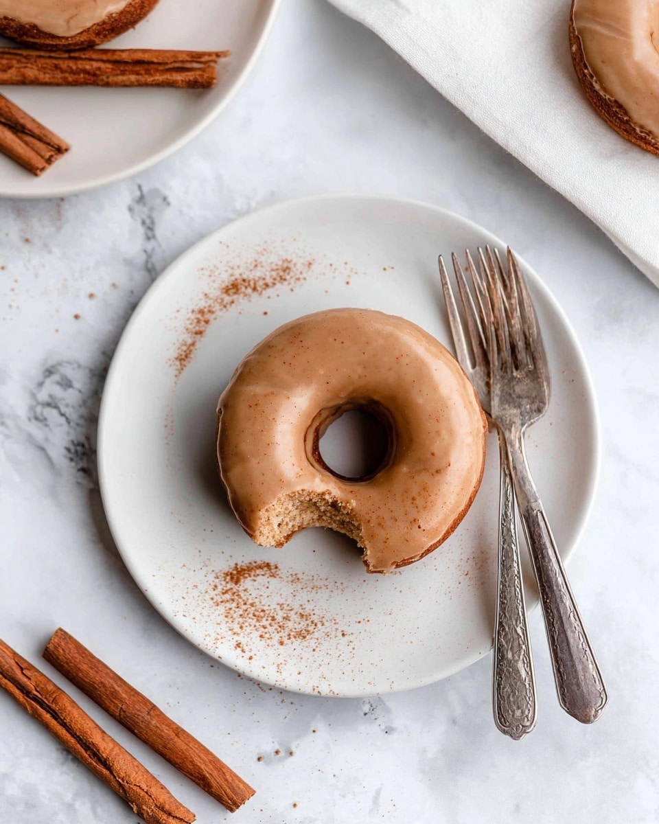 A single round donut with smooth light brown glaze covers the whole top and sides, sitting on a white plate. There is a small bite taken from the left side, revealing a soft, spongy light brown interior. Cinnamon powder is lightly sprinkled around the donut on the plate. Two silver forks with detailed handles lie beside the plate on a white marbled surface. In the top left corner, part of another white plate with a similar donut and three cinnamon sticks rests on a white cloth. More cinnamon sticks with rich brown texture sit at the bottom left on the white marbled surface. Photo taken with an iphone --ar 4:5 --v 7