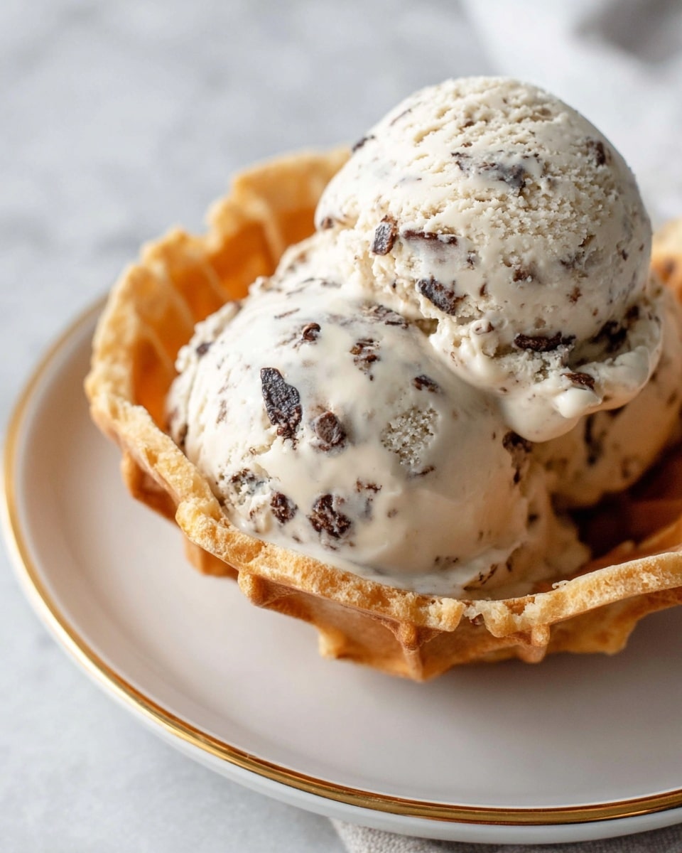 A close-up view of two scoops of light cream-colored ice cream with small dark chocolate bits mixed in, sitting inside a golden-brown waffle bowl with a slightly crispy texture. The ice cream is melting slightly, with smooth, creamy edges dripping over the sides of the scoops. The waffle bowl is placed on a white plate with a thin gold rim, set on a white marbled texture surface. photo taken with an iphone --ar 4:5 --v 7