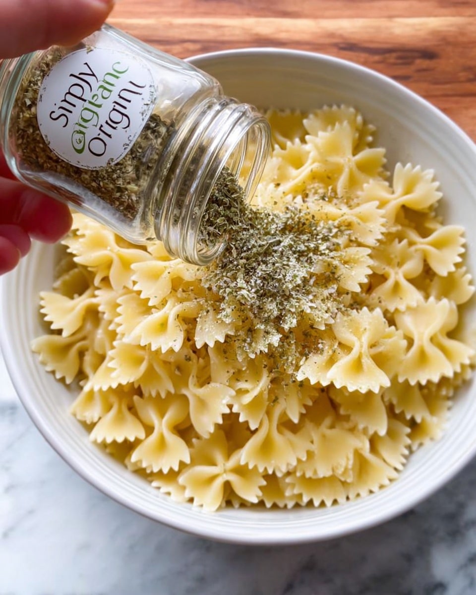 A white bowl filled with cooked farfalle pasta that is pale yellow and has a smooth texture with ridged edges. A woman's hand is holding a clear glass spice jar labeled
