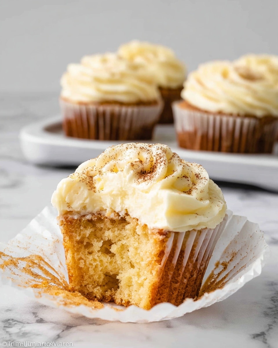 The image shows a close-up of a single cupcake with a bitten side, revealing a moist, light yellow inside. The cupcake has one layer of soft cake topped with a thick layer of creamy white frosting that is swirled smoothly and sprinkled with a light dusting of brown powder. The cupcake sits in an opened white paper wrapper, which lies on a white marbled surface. In the background, a tray holds three more cupcakes, all with the same frosting style. photo taken with an iphone --ar 4:5 --v 7