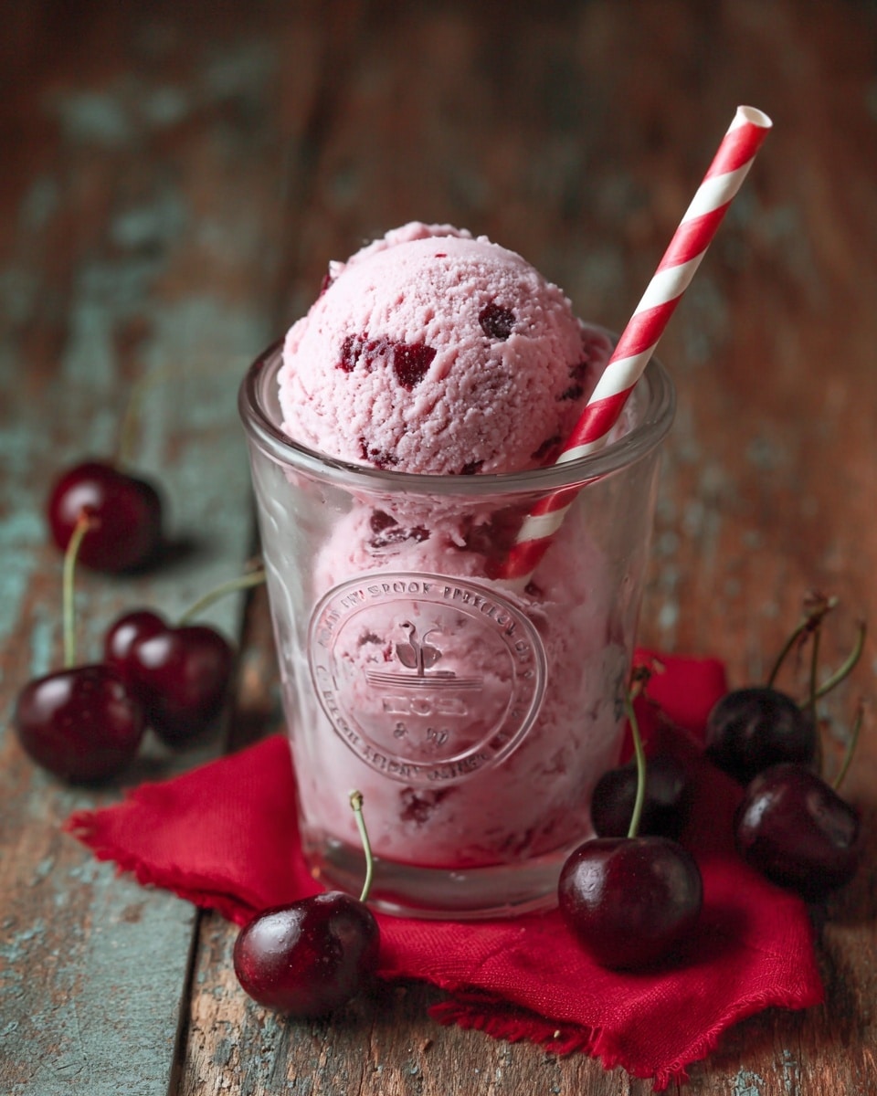 A single scoop of pink ice cream with visible cherry pieces is placed inside a clear glass with a round embossed logo on the front. A red and white striped straw leans against the glass, which sits on a red cloth on a surface resembling rustic brown wood. Several whole dark red cherries with green stems are scattered around the glass. The ice cream has a creamy, slightly textured surface with some icy crystals. Photo taken with an iphone --ar 4:5 --v 7