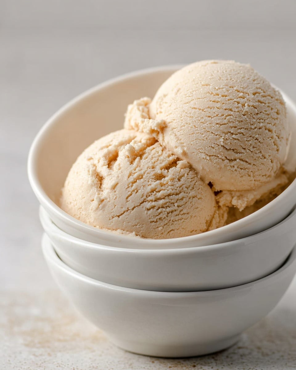 Two scoops of light beige ice cream with a smooth, slightly grainy texture sit inside a white bowl which is stacked on another white bowl, all placed on a white marbled surface. The ice cream scoops are rounded with visible soft ridges and small crumbly bits, showing a creamy consistency. The background is blurred and neutral, putting focus on the ice cream and the bowls. Photo taken with an iphone --ar 4:5 --v 7