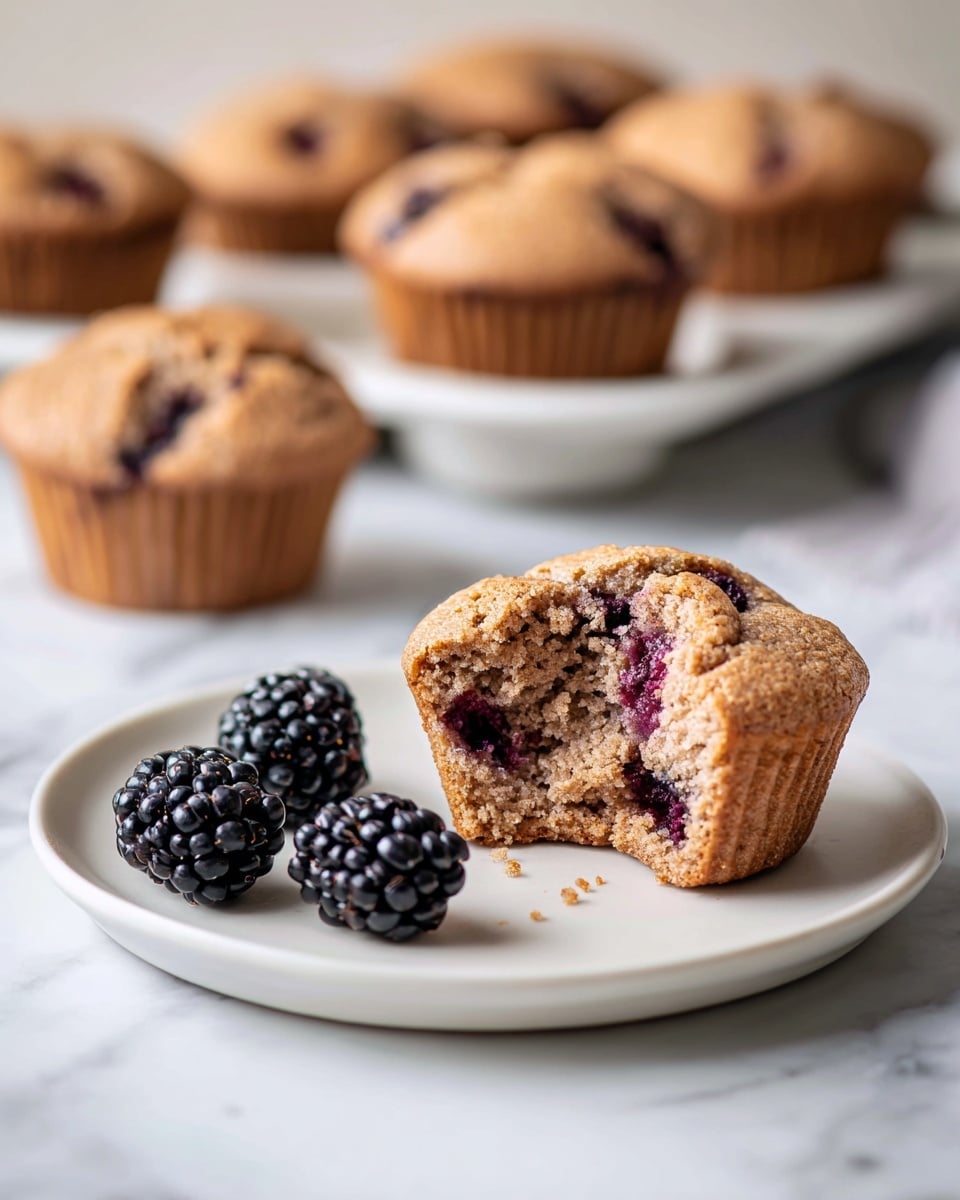 A broken open muffin sits in the middle of a white plate, showing a soft, light beige inside filled with dark purple blackberries. The muffin's top and edges have a slightly golden brown crust, with blackberries peeking through the surface, some juice staining the crumb. Three whole blackberries lie neatly in front of the muffin on the plate. In the blurry background, more muffins rest on a white tray. The plate is placed on a white marbled surface. Photo taken with an iphone --ar 4:5 --v 7