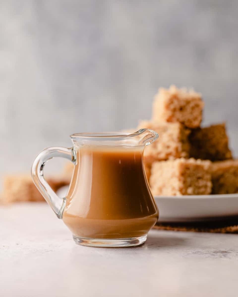 A small clear glass pitcher filled with thick light brown sauce stands on a white marbled surface. Behind it, there is a white plate holding several stacked light brown crumbly squares of dessert. The background is softly blurred with a smooth grey tone. The scene is simple and bright, highlighting the creamy texture of the sauce and the crumbly texture of the dessert. Photo taken with an iphone --ar 4:5 --v 7