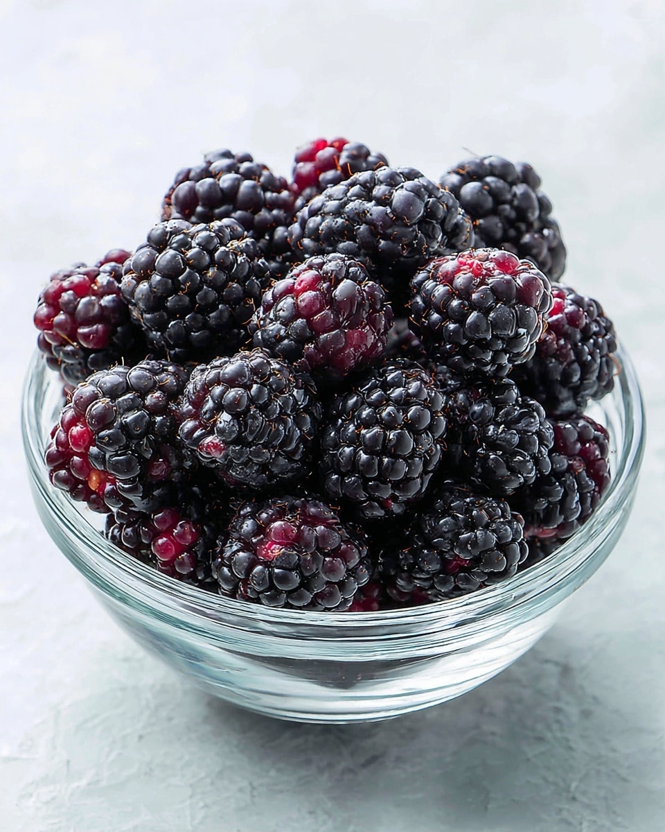 A clear glass bowl filled with many blackberries, each blackberry showing a bumpy texture and deep dark purple to black color with a few small red spots. The bowl sits on a white marbled textured surface, and the blackberries are piled up evenly, filling the bowl almost to the top. The image is bright and focuses closely on the fruit, showing the details of the blackberry skin and seeds photo taken with an iphone --ar 4:5 --v 7