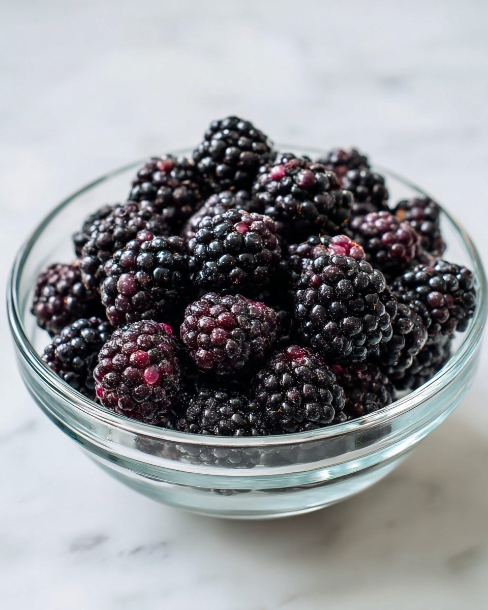 A clear glass bowl filled with many dark purple-black blackberries mixed with small white and light pink clumps that look like sugar or flour. The bowl is placed on a white marbled surface. The blackberries are piled up evenly inside the bowl, showing their bumpy texture and slightly shiny skin. photo taken with an iphone --ar 4:5 --v 7
