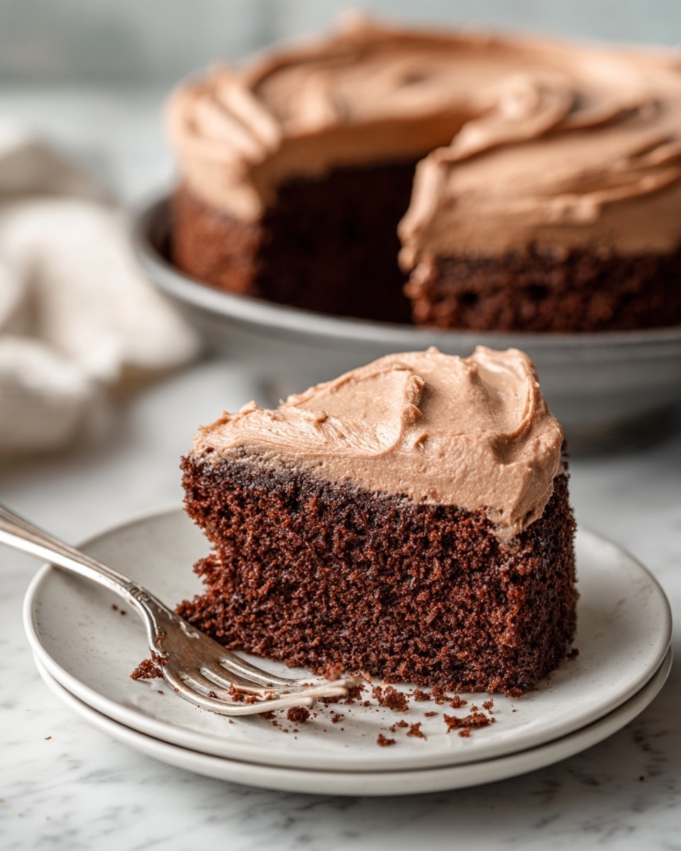 A piece of dark brown chocolate cake with a thick, smooth layer of light brown chocolate frosting on top sits on a white plate with a raised edge. The cake looks moist and soft with a slightly crumbly texture, and a bite has been taken out to show the inside. Next to the cake piece, a silver fork holds a small chunk of the cake with some frosting. In the background, there is a large square metal baking pan filled with more chocolate cake, resting on a surface with a white marbled texture. photo taken with an iphone --ar 4:5 --v 7