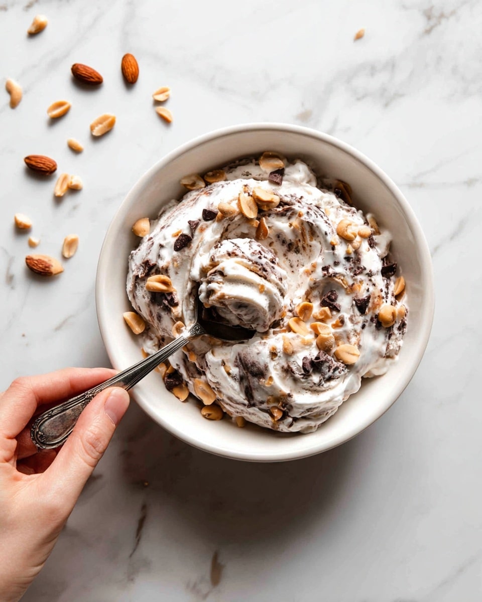 A white ramekin cup holds a creamy beige dessert that appears soft and smooth. Scattered on top and mixed inside are pieces of dark brown chocolate chunks and light brown chopped peanuts, adding texture with their rough and uneven shapes. A vintage silver spoon inside the cup lifts a scoop of the creamy dessert with the chocolate and peanuts mixed in. There is a woman's hand holding the spoon, visible on the left side of the image. The background is a clean white marbled surface with a few chocolate and peanut pieces scattered around, giving the scene a fresh and simple look. Photo taken with an iphone --ar 4:5 --v 7