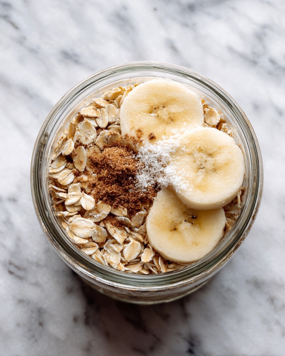 A clear glass blender cup on a white marbled surface holds layers of light tan rolled oats filling most of the container, topped with two thick banana slices of pale yellow color. On top of the oats and banana slices, there is a small pile of medium brown powder and a sprinkle of white granules. The view is from above, showing the textures of oats, the soft smooth banana, and the powders clearly. Photo taken with an iphone --ar 4:5 --v 7