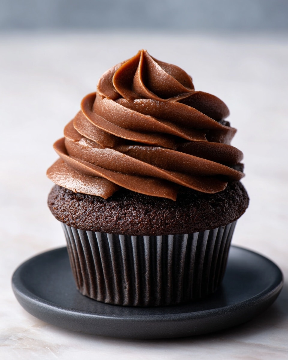 A single chocolate cupcake sits in a brown paper liner as the base layer, showing a moist and slightly textured surface. On top of this base, a large, thick swirl of rich, creamy dark chocolate frosting is piped in multiple waves, creating a tall, fluffy peak with soft ridges and folds. The cupcake is placed on a dark plate with a matte finish, all set against a white marbled textured background. photo taken with an iphone --ar 4:5 --v 7